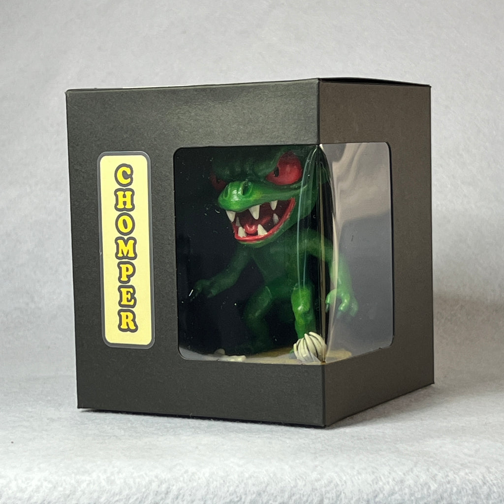 Green toy dinosaur figure named 'Chomper' in a black box packaging on a gray background