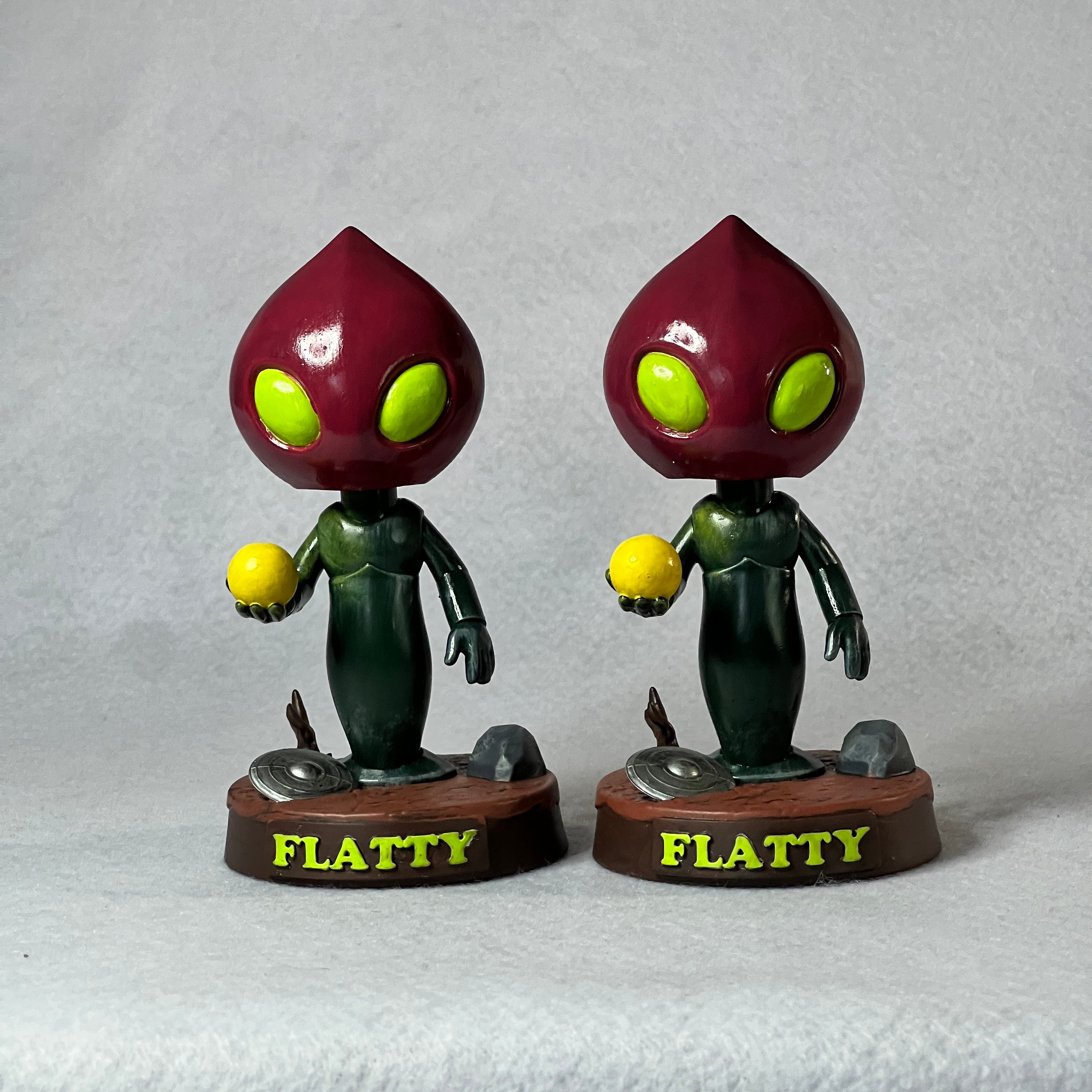 Two identical figurines of a character named Flatty on a gray background