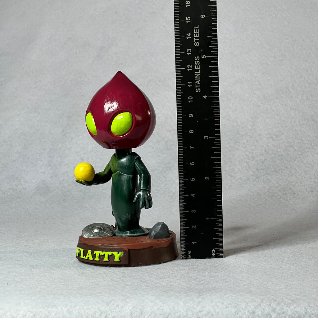Flattie figurine holding a yellow ball next to a ruler on a gray background