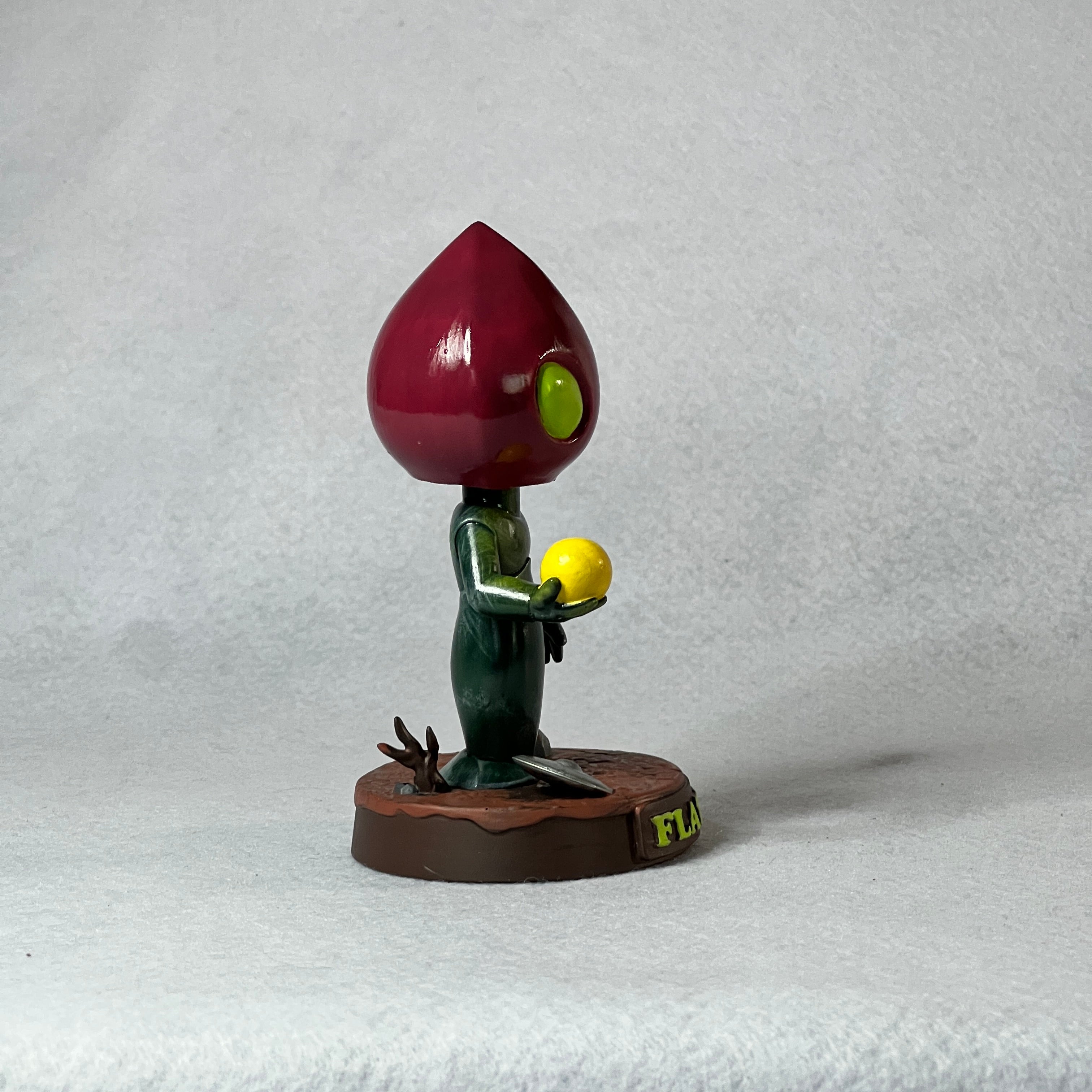 Figurine with a large red head and green body holding a yellow object on a white background