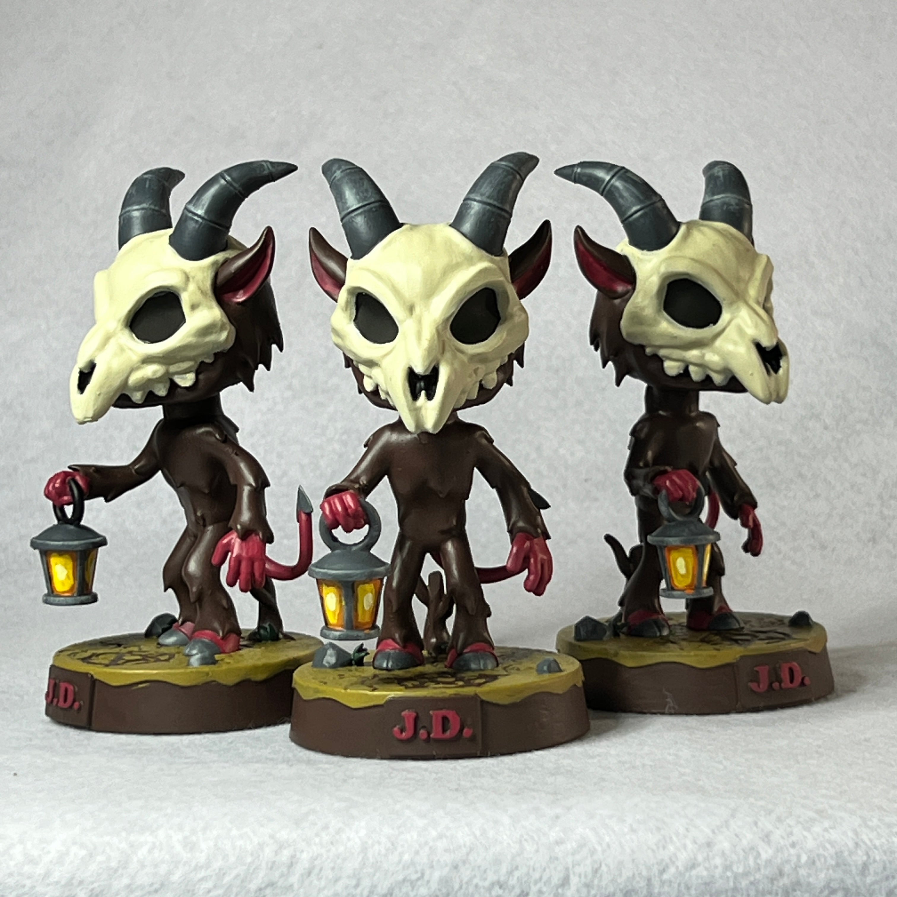 Three figurines of demon skulls with brown bodies, red hands and feet, and black horns on a gray background.