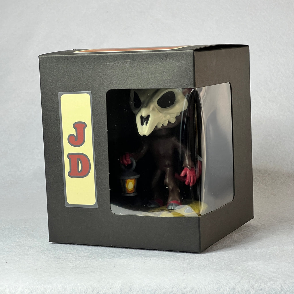 Skull-shaped figurine in a black box with 'JD' label on a light gray background