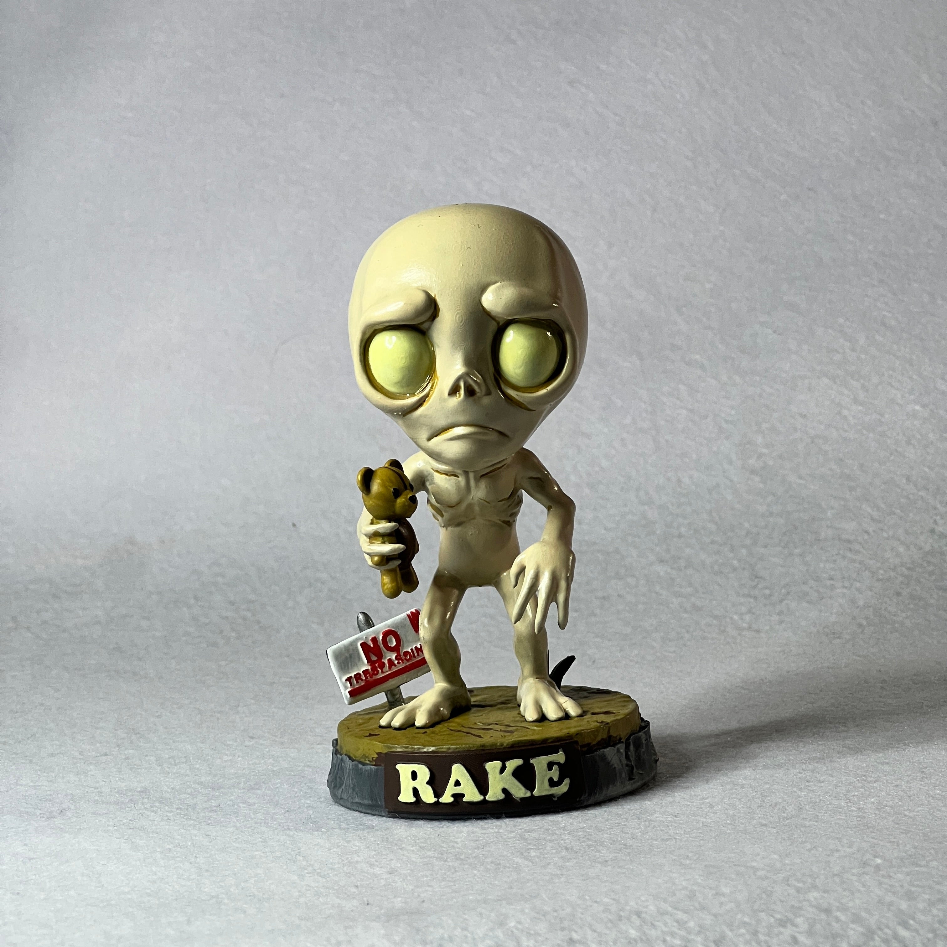 Alien figurine holding a teddy bear and a sign with text on a gray background