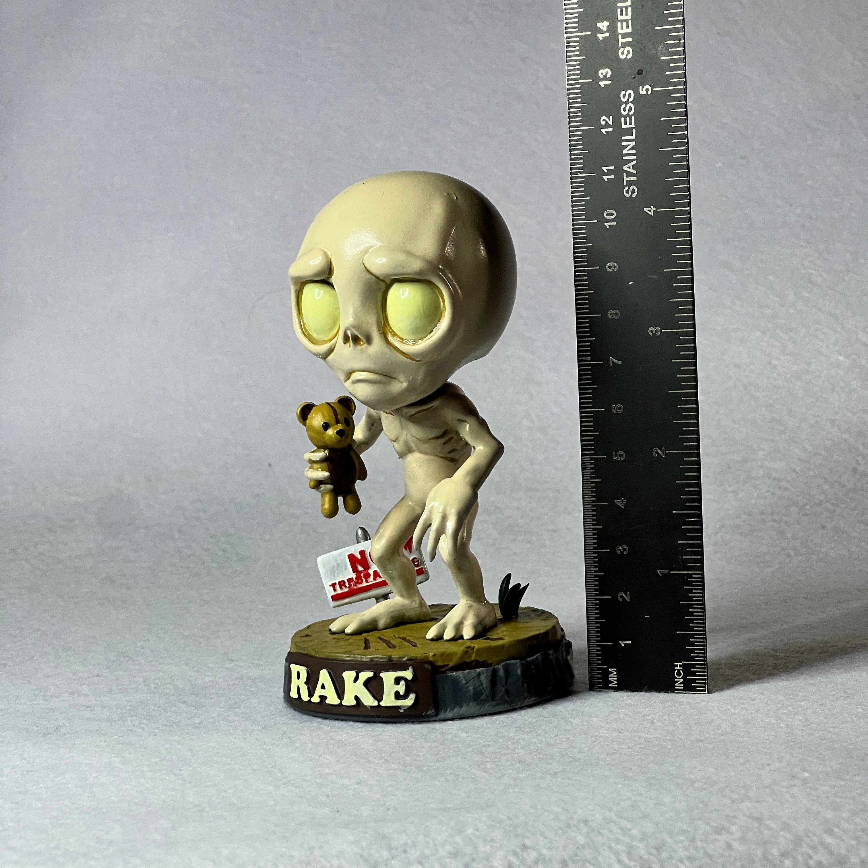 Alien figurine holding a teddy bear with a 'Nope' sign, next to a ruler for scale, on a gray background.