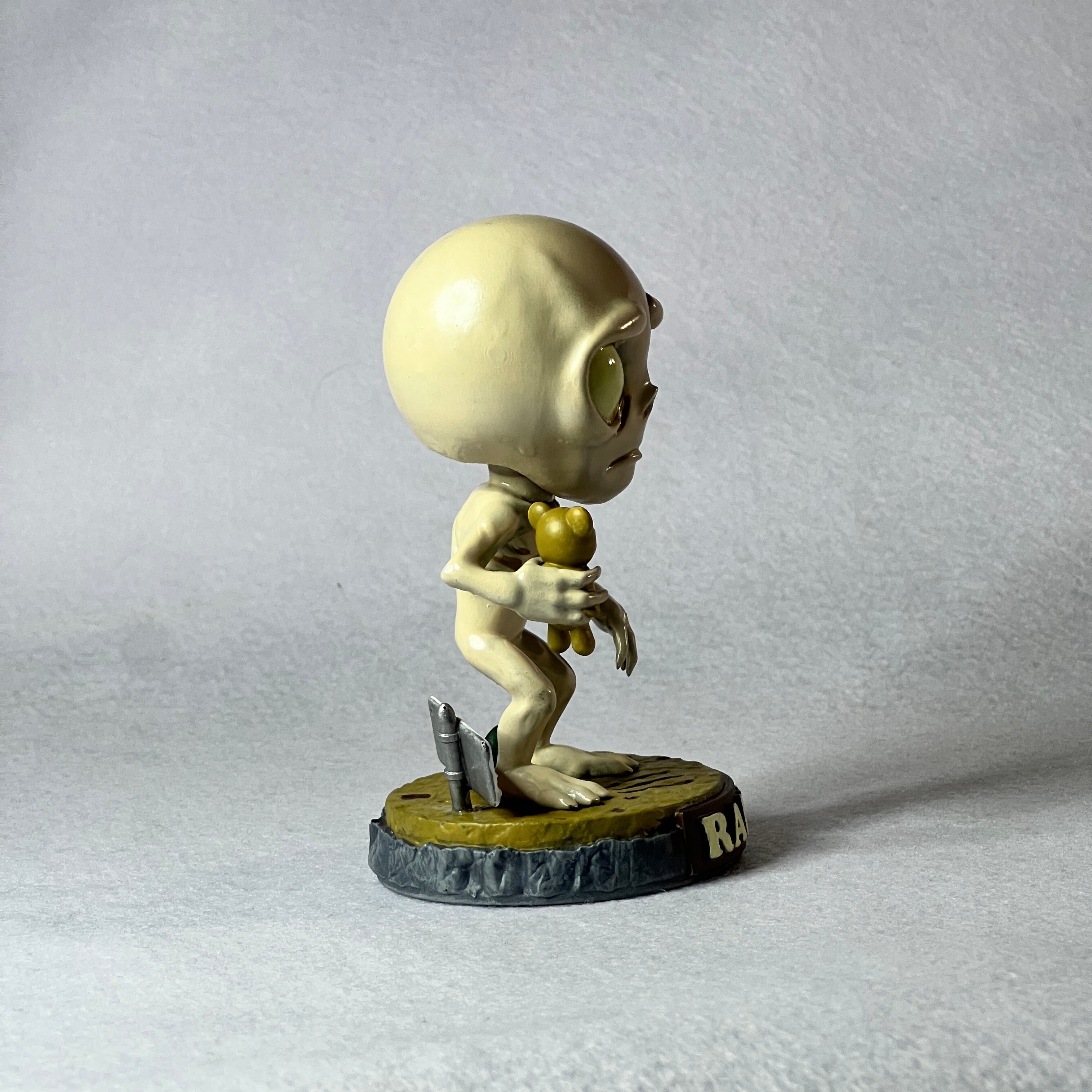 Small figurine of a humanoid with a round head and limbs, holding an apple, on a textured gray background.