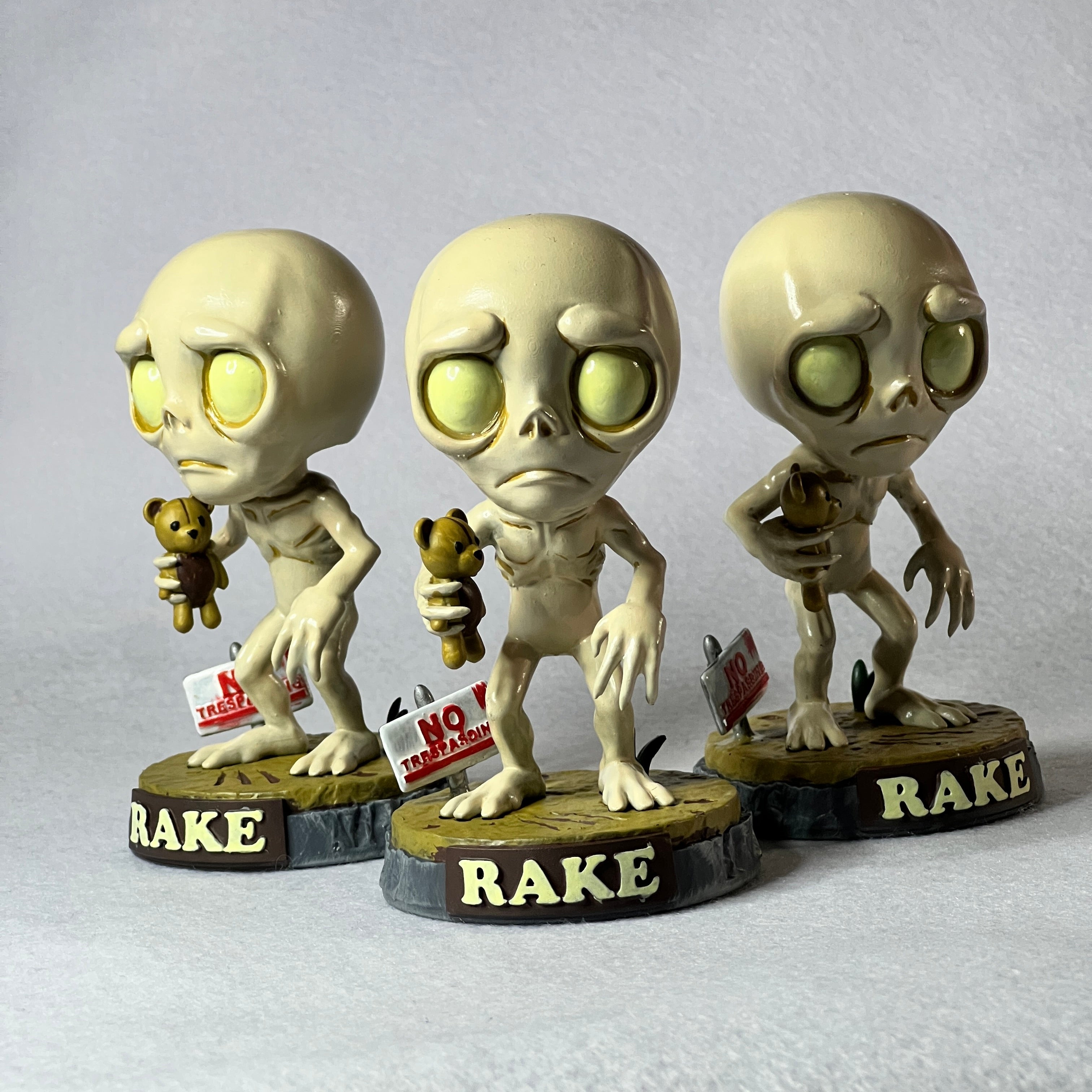 Three alien figurines holding teddy bears and signs with text on a gray background
